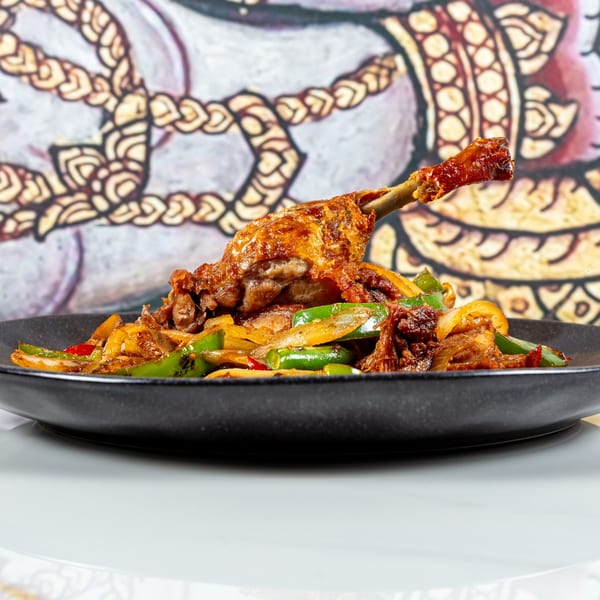 Crispy duck leg with green peppers and onions on a black plate, against a Thai art background.