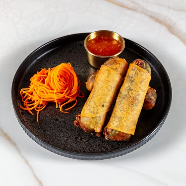 Two crispy Thai spring rolls with shredded carrots and sweet chili sauce on a black plate.