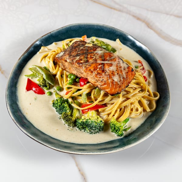 Grilled salmon fillet over linguine pasta with creamy sauce, broccoli, peas, and red bell peppers.
